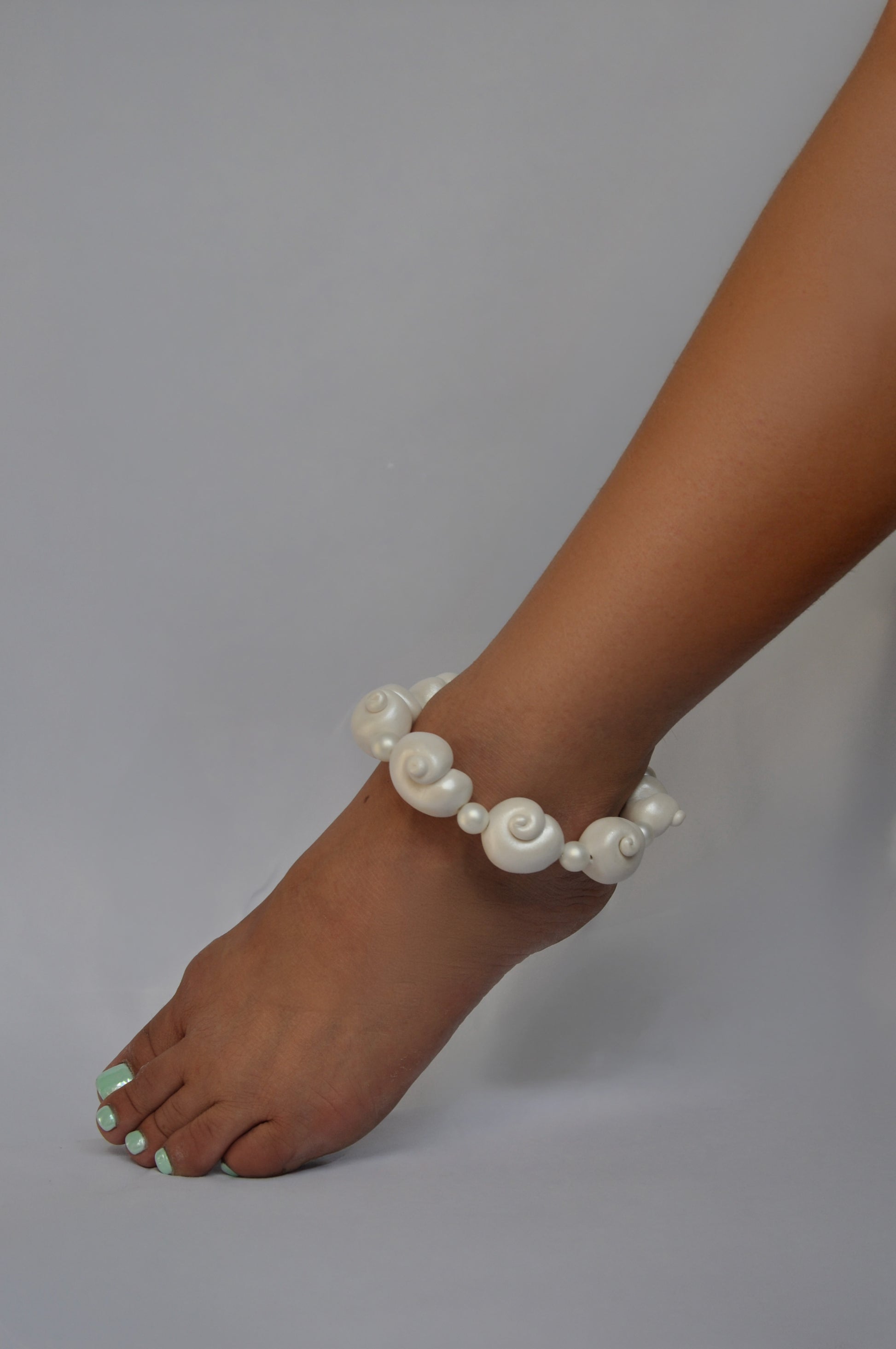 A white beaded anklet with spiral shell-like beads worn on a person's ankle, shown against a plain light gray background.